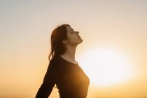 Side view of happy female standing with eyes closed on hill and enjoying freedom while admiring mountainous scenery at sunset — Stock Photo