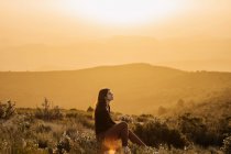 Vista lateral do viajante feminino tranquilo sentado na colina com os olhos fechados e desfrutando da natureza nas terras altas ao pôr do sol — Fotografia de Stock