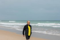 Desportista idoso caminhando na praia perto do mar acenando durante o treino de fitness no verão — Fotografia de Stock