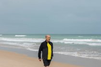 Anciano deportista caminando en la playa cerca del mar ondeando durante el entrenamiento de fitness en verano - foto de stock