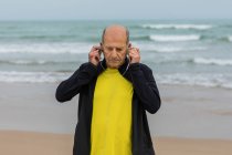 Älterer männlicher Athlet setzt Kopfhörer auf, während er sich auf Fitnesstraining am Strand in der Nähe des winkenden Meeres vorbereitet — Stockfoto