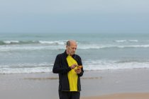 Atleta masculino envejecido escuchando música en auriculares y usando un teléfono inteligente durante el entrenamiento en la playa cerca del mar ondulante - foto de stock