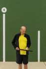 Пожилой спортсмен с теннисным мячом и ракеткой смотрит вниз, стоя у зеленой стены во время тренировки в тренажерном зале — стоковое фото