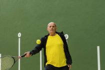 Пожилой спортсмен бьет по мячу ракеткой, играя в теннис против зеленой стены в спортзале — стоковое фото