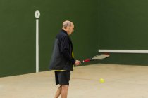 Idosos atleta masculino batendo bola com raquete enquanto joga tênis contra a parede verde no ginásio — Fotografia de Stock