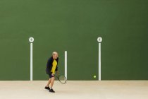 Idosos atleta masculino batendo bola com raquete enquanto joga tênis contra a parede verde no ginásio — Fotografia de Stock