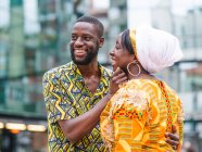 Side view of cheerful black couple in bright outfit with ornament embracing while standing on urban pavement looking away — Stock Photo