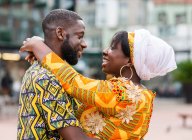 Side view of cheerful black couple in bright outfit with ornament embracing while standing on urban pavement and looking at each other — Stock Photo