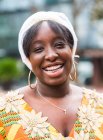 Fröhliche junge Afrikanerin in hellem Gewand mit floralem Ornament blickt in die Kamera, die in der Stadt steht — Stockfoto