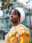 Vue latérale de la jeune femme africaine en tenue lumineuse avec ornement floral regardant la caméra debout dans la ville — Photo de stock