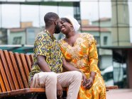 Allegro donna africana in abito tradizionale seduto vicino al fidanzato rasato in abiti ornamentali sulla panchina della città — Foto stock