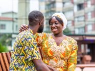 Весела африканка в традиційному одязі сидить поруч неголеного хлопця в декоративному одязі на міському лаві. — стокове фото