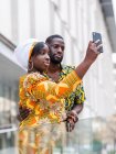 From below of content African woman in colorful apparel with ornament near cheerful boyfriend taking self portrait on cellphone in town — Stock Photo