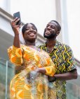 From below of content African woman in colorful apparel with ornament near cheerful boyfriend taking self portrait on cellphone in town — Stock Photo