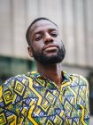 Thoughtful unshaven African American male in ornamental shirt looking at camera while standing in summer city — Stock Photo