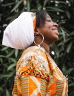 Visão lateral da jovem fêmea africana alegre com aparelho no lenço com olhos fechados contra a planta no verão — Fotografia de Stock