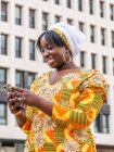 Знизу усміхненої чорної жінки в декоративному одязі, що переглядає мобільний телефон, стоячи в місті — стокове фото