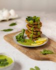 Свежие вкусные травяные оладьи, сложенные на тарелке и украшенные зеленым соусом и ломтиком лимона, подаваемые на деревянной доске — стоковое фото