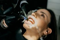 Обрізати нерозпізнаний стоматолог, вишиваючи фоточутливу пасту, лікуючи зуби жінки з екстрактором слини та рефрактором у роті — стокове фото