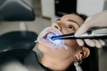 Crop dentista irreconhecível usando luz de cura dental durante o tratamento de dentes de fêmea com ejetor de saliva e retrator na boca — Fotografia de Stock