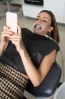 Пациентка с ретрактором во рту лежит на стоматологическом стуле и делает самоснимок на смартфоне перед лечением в клинике — стоковое фото