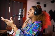 Vue latérale de la jeune chanteuse afro-américaine dans un casque enregistrant une chanson contre un producteur de musique souriant en studio — Photo de stock