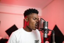 Jovem vocalista afro-americano com boca aberta e corte de cabelo moderno cantando em microfone profissional — Fotografia de Stock