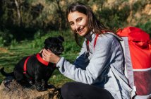 Side view of cheerful young woman with backpack caressing cute little dog while resting during hiking in forest — Stock Photo