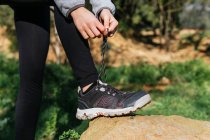 Vista lateral de la cosecha irreconocible mujer excursionista atando cordones en zapatillas de deporte durante el descanso durante la aventura de verano en el bosque - foto de stock
