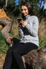 Позитивная молодая туристка в активной одежде сидит на стволе дерева и проверяет маршрут на мобильном телефоне, проводя солнечный день в зеленом лесу — стоковое фото