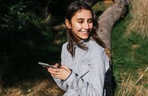 Positive junge Wanderin in Aktivkleidung sitzt auf einem Baumstamm und checkt mit dem Handy die Route, während sie einen sonnigen Tag im grünen Wald verbringt und wegschaut — Stockfoto