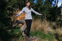 Повне тіло щасливої молодої жінки в спортивному одязі балансує на стовбурі падіння дерева під час прогулянок по зеленому лісу в сонячний день — стокове фото