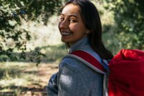 Вид ззаду щасливої оптимістичної молодої жінки-хокеїстки в активному одязі з рюкзаком, насолоджуючись подорожжю в зеленому лісі в сонячний день — стокове фото