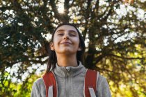 Glücklich optimistische junge Wanderin in Aktivkleidung mit Rucksack genießt Fahrt im grünen Wald bei sonnigem Tag mit geschlossenen Augen — Stockfoto