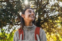 Glücklich optimistische junge Wanderin in Aktivkleidung mit Rucksack genießt Fahrt im grünen Wald bei sonnigem Tag — Stockfoto