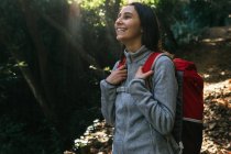 Happy optimistic young female hiker in activewear with backpack enjoying journey in green forest in sunny day — Stock Photo