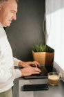 Вид сбоку улыбающегося мужчины средних лет, работающего на прилавке с нетбуком и чашкой кофе на кухне утром — стоковое фото