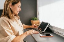 Seitenansicht einer reifen Frau, die am Tresen in der Küche sitzt und morgens auf dem Tablet surft — Stockfoto