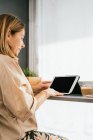 Side view of mature female sitting at counter in kitchen and browsing tablet in morning — Stock Photo