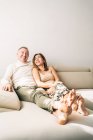 Délicieux couple d'âge moyen assis sur un canapé confortable dans le salon à la maison tout en souriant et profiter week-end ensemble — Photo de stock