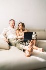 Mediana edad sonriente pareja abrazándose en el sofá en la sala de estar y viendo la película en netbook mientras se relaja en casa en fin de semana - foto de stock