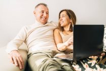 У середньому віці усміхнена пара присідає на дивані у вітальні і дивиться фільм на нетбуці, відпочиваючи вдома на вихідних — стокове фото