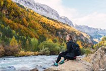 Vista lateral del excursionista femenino con mochila sentada en roca cerca del río en las montañas de los Pirineos mientras disfruta de vacaciones en el Parque Nacional Ordesa y Monte Perdido - foto de stock