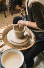 Vista lateral de una artesana irreconocible enfocada usando una rueda de cerámica y creando loza hecha a mano en el taller - foto de stock