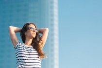 Baixo ângulo de alegre despreocupado jovem fêmea em óculos de sol vestindo camisa listrada casual e jeans desfrutando de dia ensolarado de verão — Fotografia de Stock