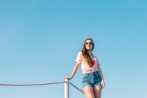 Снизу современной женщины тысячелетия в солнцезащитных очках в джинсовых шортах и стильной повседневной рубашке, смотрящей в сторону, стоя на фоне голубого неба в летний день — стоковое фото