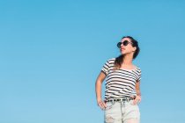 Bajo ángulo de alegre mujer joven sin preocupaciones en gafas de sol con camiseta de rayas casuales y vaqueros disfrutando de un día soleado de verano - foto de stock