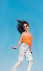 From below full body of modern hipster female in sunglasses wearing shirt and stylish ripped jeans jumping high against blue sky — Stock Photo