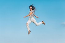 From below full body of modern hipster female in sunglasses wearing striped shirt and stylish ripped jeans jumping high against blue sky — Stock Photo