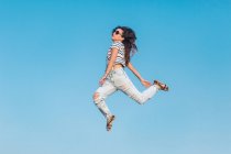 Du bas du corps complet de la femme hipster moderne en lunettes de soleil portant une chemise rayée et un jean déchiré élégant sautant haut contre le ciel bleu — Photo de stock